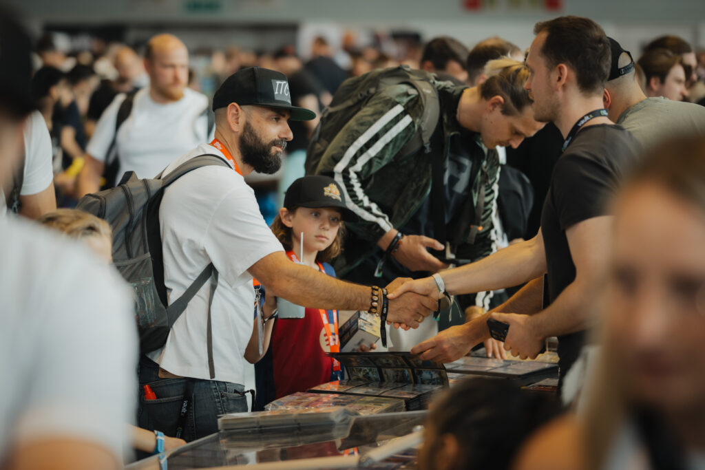 Collectors shaking hands across a dealer table in the busy CARDMADNESS hall